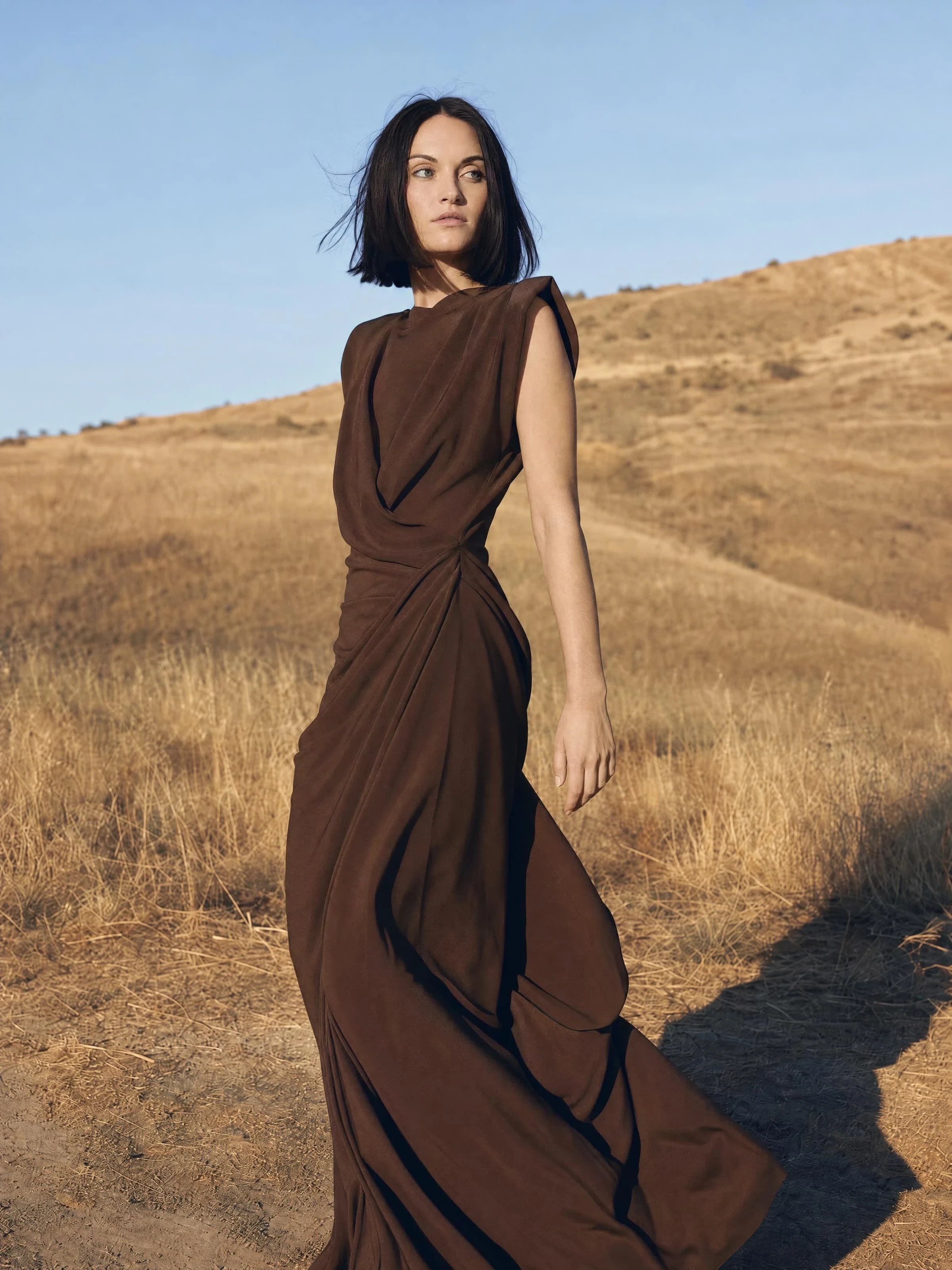 A model in a draped chocolate dress on sunlit golden hills, hair moving in the wind