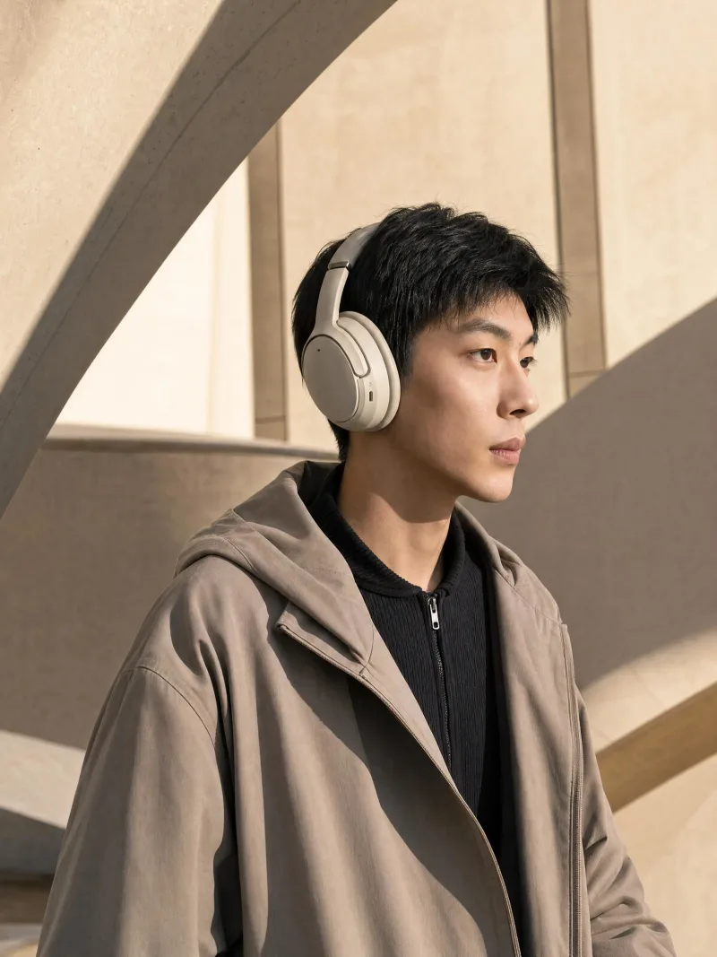 Danny in a stone-coloured hooded coat wearing cream over-ear headphones under a sunlit concrete arch